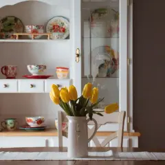 Stary kredens w salonie, pełen kolorowej porcelany i ozdobnych talerzy. Na stole bukiet żółtych tulipanów w dzbanku.