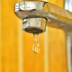 Kropla wody wisi na kranie, który źle miesza wodę. Tło jest w kolorze żółtym.