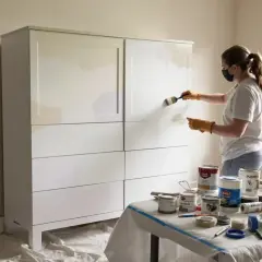 Kobieta w masce i rękawiczkach maluje białą szafę pędzlem. Widać puszki z farbą i taśmę. To instrukcja, jak pomalować szafę.