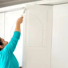 Kobieta maluje białą szafę przesuwną wałkiem. Odświeża mebel, by wyglądał jak nowy.