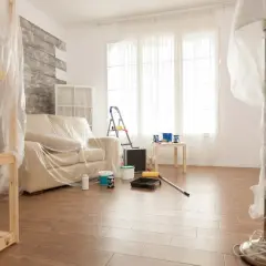 Przygotowania do malowania paneli podłogowych. Meble i lampa przykryte folią, na podłodze wałek i kuweta z farbą.