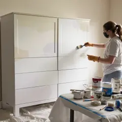 Kobieta w masce i rękawiczkach maluje białą szafę. Na stole obok puszki z farbą i pędzle.