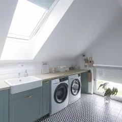 Jasna pralnia na poddaszu ze skosami, z pralką, suszarką, dużą wanną i oknem dachowym.
