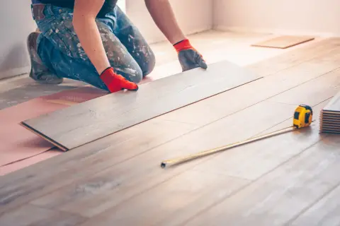 Ręce w rękawiczkach układają panele, tworząc podłogę pływającą. Miarka leży obok.