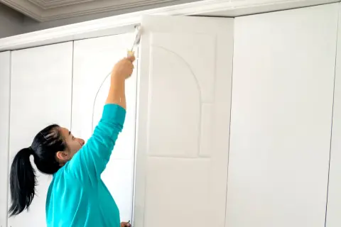 Kobieta maluje białą szafę przesuwną wałkiem. Odświeża mebel, by wyglądał jak nowy.