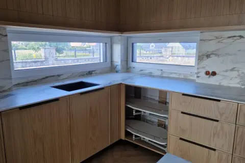 Nowoczesna kuchnia z drewnianymi frontami i marmurowym blatem. Okna wychodzą na ogród, a parapet z blatu kuchennego tworzy spójną całość.