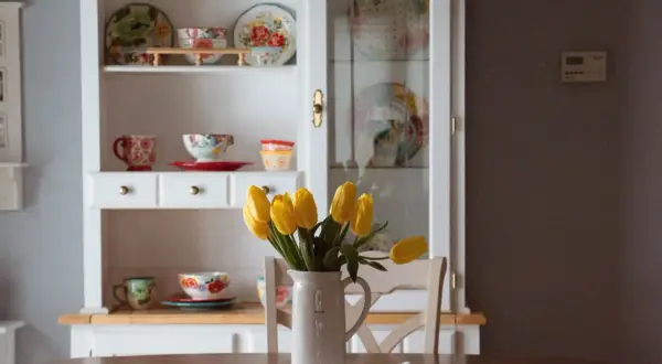 Stary kredens w salonie, pełen kolorowej porcelany i ozdobnych talerzy. Na stole bukiet żółtych tulipanów w dzbanku.