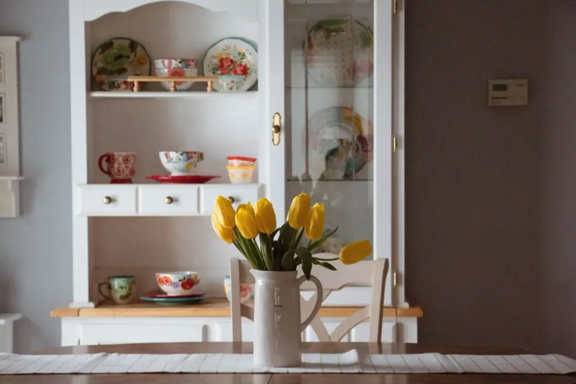 Stary kredens w salonie, pełen kolorowej porcelany i ozdobnych talerzy. Na stole bukiet żółtych tulipanów w dzbanku.