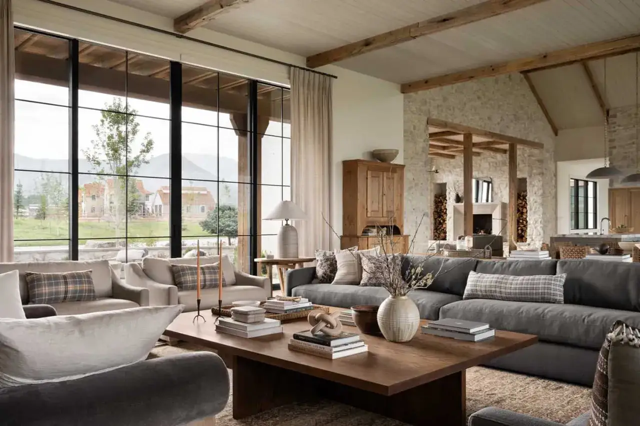 Przytulny salon w stylu farmhouse z kamiennym kominkiem, drewnianymi belkami i dużymi oknami z widokiem na g&oacute;ry.