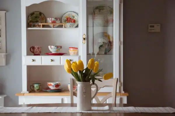 Stary kredens w salonie, pełen kolorowej porcelany i ozdobnych talerzy. Na stole bukiet żółtych tulipanów w dzbanku.